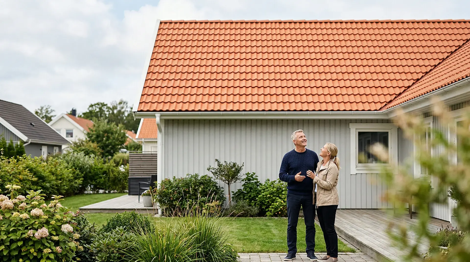 Par står utanför hus med rött tegeltak och välskött trädgård.