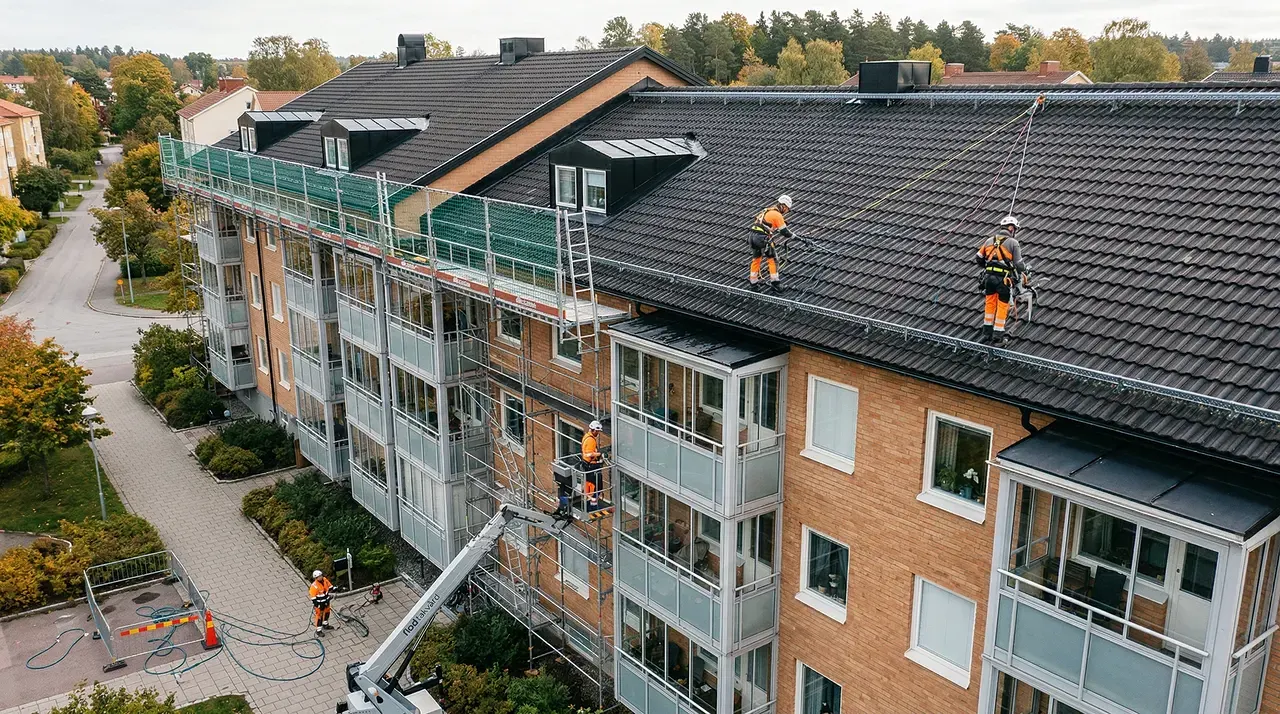 Takarbetare i orange skyddskläder renoverar flervåningshus med scaffolding och lift.